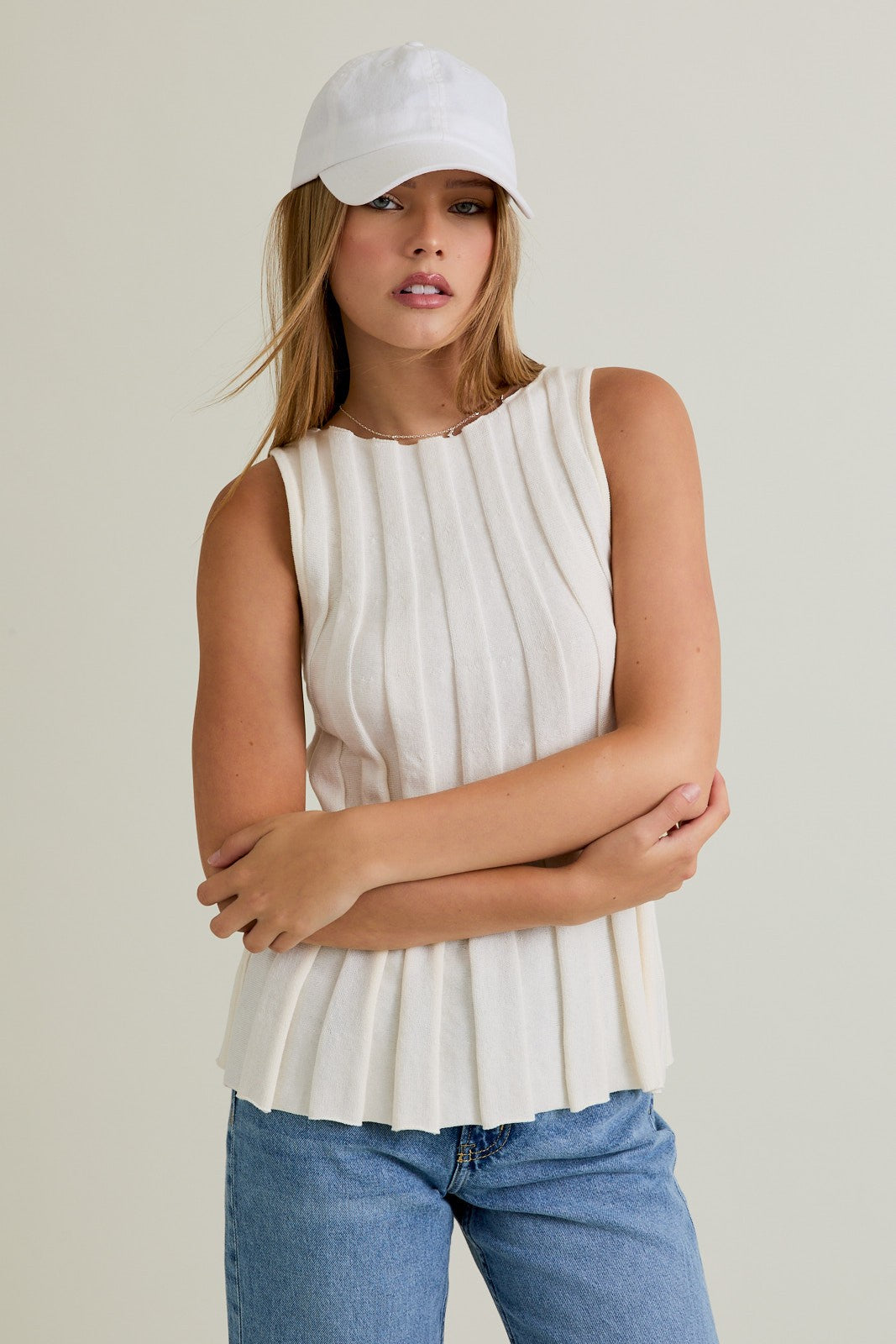 Woman wearing a white sleeveless top and blue jeans with a white cap on a plain background